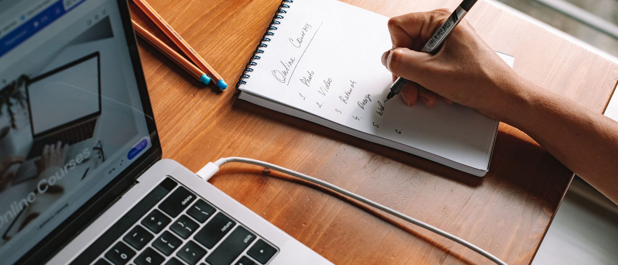 A person taking handwritten notes beside an open laptop on a wooden desk